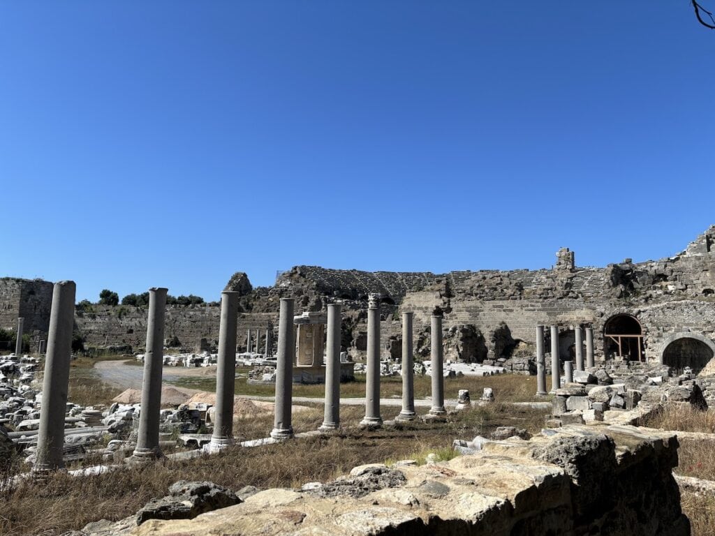 Zu den antiken Ruinen in der Altstadt von Side zählen mehrere stehende Säulen und teilweise eingestürzte Steinstrukturen unter einem klaren blauen Himmel.