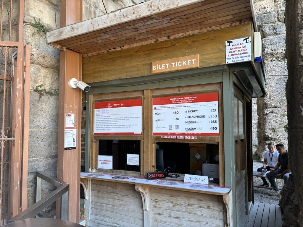 Ticketschalter aus Holz, im bezaubernden Außenbereich der Altstadt Side gelegen, mit Preisen in türkischer Lira für Eintritt, Headset-Verleih und Museumszugang.