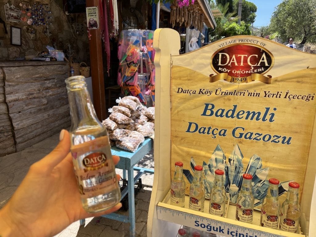 Eine Person hält eine kleine Flasche Datça-Mandellimonade in der Hand, im Hintergrund ein Regal mit ähnlichen Flaschen und türkische Mandeln.