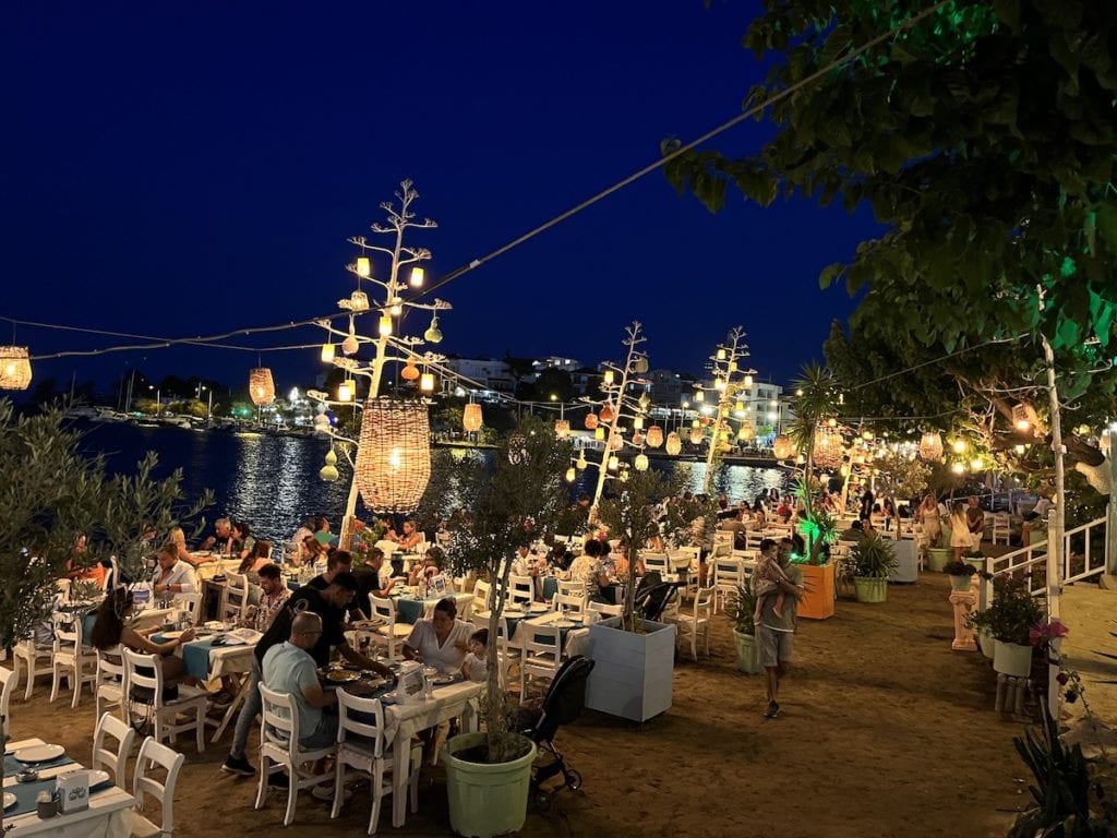 Abendessen im Freien in Datca am Wasser, mit Gästen an Tischen, die von Lichterketten und Laternen beleuchtet werden.