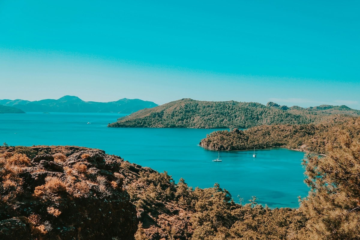 Eine ruhige Küstenszene in Datca mit üppigem Grün und klarem blauen Wasser, die eine ruhige Bucht mit Booten und sanften Hügeln in der Ferne zeigt.
