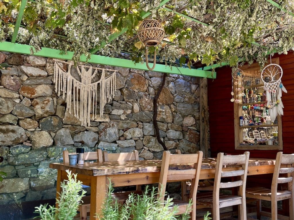 Rustikaler Essbereich im Freien in Datca mit einer Steinmauer, Holztischen und dekorativen Hängegegenständen unter einer mit Weinreben bewachsenen Pergola.