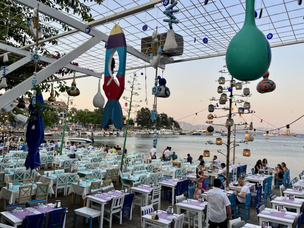 Essbereich im Freien am Meer in Datca mit farbenfrohen hängenden Ornamenten und Gästen, die ihre Mahlzeiten bei Sonnenuntergang genießen.