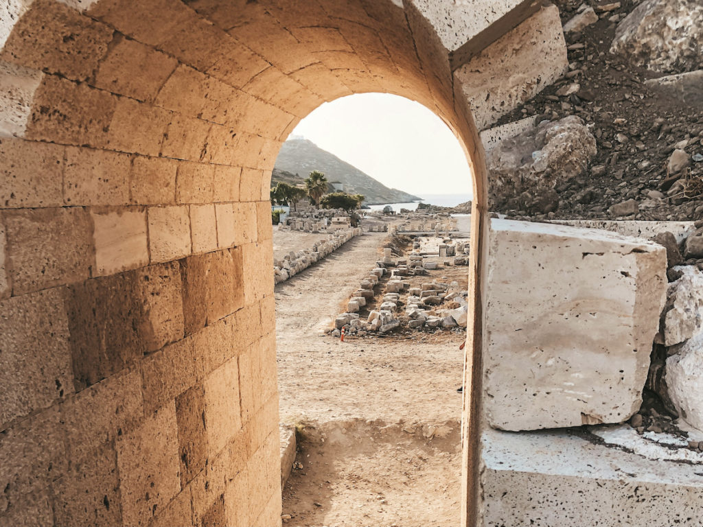 Ein steinerner Torbogen umrahmt den Blick auf die antike archäologische Stätte Datca mit Ruinen und Bergen in der Ferne.