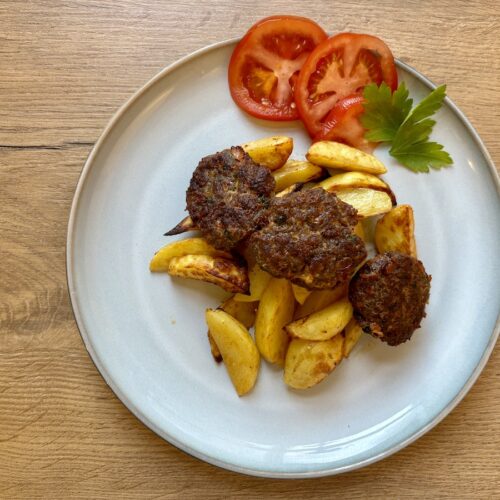 Ein Teller mit türkischen Fleischbällchen und Kartoffelecken, garniert mit Tomatenscheiben und Petersilie, auf einem Holztisch.