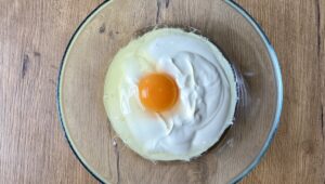 Joghurt mit rohem Ei mit intaktem Eigelb, umgeben von Eiweiß, in einer durchsichtigen Glasschüssel auf einer Holzoberfläche.