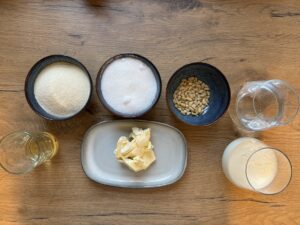 Verschiedene Zutaten auf einem Holztisch, darunter Zucker, Grieß, Butter, Pinienkerne, Wasser und Milch.