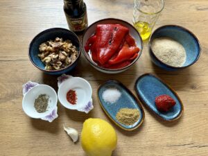 Zutaten für ein auf einem Tisch angerichtetes Gericht: Walnüsse, geröstete rote Paprika, Olivenöl, Semmelbrösel, Tomatenmark, Gewürze, eine Knoblauchzehe, eine Zitrone und eine Flasche Granatapfelmelasse