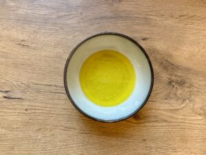 In einer Keramikschale mit weißer Innenseite ruht eine kleine Menge gelbes Öl mit Zitronensaft und Knoblauch. Die Schale steht auf einer Holzoberfläche.