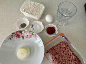 Zu den Zutaten für Manti auf einer Arbeitsplatte gehören Hackfleisch, ein Ei, ein Glas Wasser, Zwiebel, Mehl und kleine Schälchen mit Pfeffer, Salz und Tomatenmark