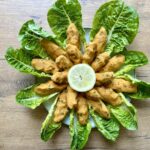 Ein Teller mit zwölf orangefarbenen Linsen-Bällchen, angeordnet auf einem Bett aus Salat, zentriert um eine Zitronenscheibe.