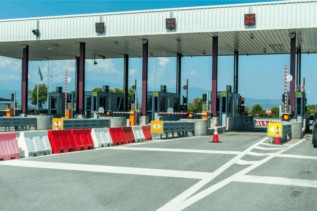 Eine Autobahnmautstelle mit mehreren Fahrspuren. Man sieht rote und weiße Schranken, Verkehrsleitkegel und Richtungspfeile. Der Himmel ist klar, im Hintergrund sieht man Berge, perfekt für alle, die „Mit dem Auto in die Türkei“ reisen möchten.