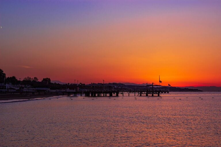 Sandstrand bei Sonnenuntergang mit ruhigem Wasser, einem ins Wasser ragenden Pier und einem von Orange und Pink zu Dunkelviolett wechselnden Himmel – eine idyllische Kulisse für einen erholsamen Spa-Aufenthalt oder eine gemütliche Runde Golf im Voyage Belek.