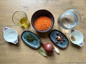 Auf einem Holztisch liegen die Zutaten für ein klassisches Mercimek-Gericht: eine Schüssel mit roten Linsen, ein Glas Wasser, eine Schüssel mit Öl, Zwiebeln, Knoblauch, Lorbeerblatt, eine Schüssel mit Haselnüssen, Schüsseln mit Gewürzen und Kräutern und eine kleine Schüssel mit Salz.