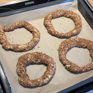 Vier rohe Teigringe, die mit Körnern umhüllt sind, werden auf ein mit Backpapier ausgelegtes Backblech gelegt und sind bereit zum Backen.