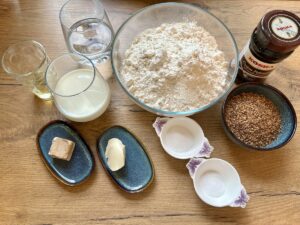 Ein Holztisch mit Backzutaten. Dazu gehören Mehl, Hefe, Salz, Zucker, geschrotete Körner, Milch, Wasser, Butter und ein Glas Öl.