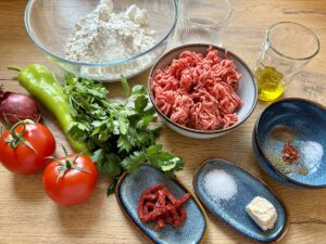 Zutaten auf einem Holztisch: Hackfleisch, Mehl, Hefe, Tomaten, Zwiebeln, grüne Paprika, Petersilie und Olivenöl. Gewürze und Tomatenmark für die Zubereitung von Lahmacun.