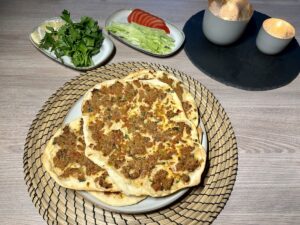 Teller mit mehreren fertigen Lahmacun stehen auf einem Holztisch, umgeben von geschnittenen Tomaten, Salat und Petersilie. Brennende Kerzen im Hintergrund.