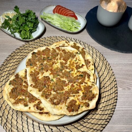 Teller mit mehreren fertigen Lahmacun stehen auf einem Holztisch, umgeben von geschnittenen Tomaten, Salat und Petersilie. Brennende Kerzen im Hintergrund.