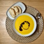 Eine Schüssel Kürbissuppe mit Kürbiskernöl, Serviert Auf Einem Gewebten Tischset. Zusammen Mit Einem Löffel Und Einem Teller und geschnittenem Brot.