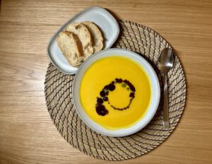 Eine Schüssel Kürbissuppe mit Kürbiskernöl, Serviert Auf Einem Gewebten Tischset. Zusammen Mit Einem Löffel Und Einem Teller und geschnittenem Brot.