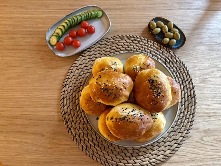 Ein Teller türkischen Pogaca mit Kartoffelfüllung Auf Einer Geflochtenen Matte Wird Von Einer Kunstvoll Arrangierten Beilage Aus Gurkenscheiben, Kirschtomaten Und Einer Kleinen Schale Oliven Begleitet.