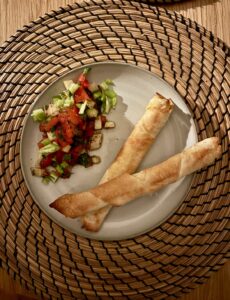 Ein Teller Mit Zwei Knusprig Gerollten Sigarabörek Und Einer Beilage Aus Gavurdağı Salatası, Einem Mit Frisch Geschnittenem Gemüse Gefüllten Gericht, Steht Auf Einem Gewebten Tischset.