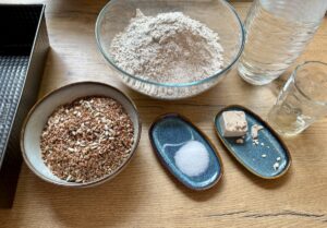 Schüsseln und teller mit mehl, körnermischung, salz, frischer hefe, Apfelessig und wasser stehen auf einem holztisch neben einer backform – perfekt für die zubereitung von selbstgemachtem dinkel vollkornbrot.