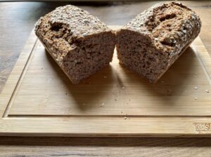 Auf einem hölzernen schneidebrett liegt ein halbierter laib dinkel vollkornbrot, darüber sind einige krümel und haferflocken verstreut.