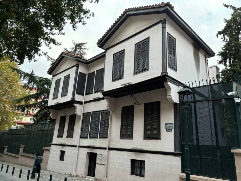 Ein zweistöckiges weißes Gebäude mit schwarzen Fensterläden, einem Ziegeldach und einem Straßenschild an der Außenwand steht in Thessaloniki in Griechenland, umgeben von Bäumen, einem Zaun.