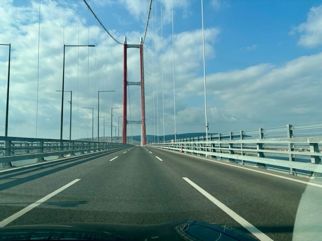 Blick aus einem E Auto auf eine Hängebrücke mit roten Türmen in der Türkei, mit mehreren Fahrspuren, Metallgeländern und Straßenlaternen unter einem teilweise bewölkten blauen Himmel.