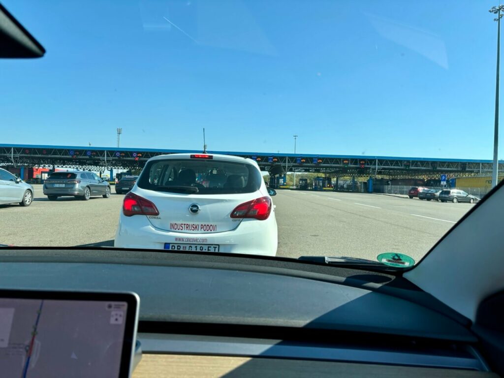 Blick aus dem Inneren eines Autos, das sich einer Mautstelle in Serbien nähert, mit einem weißen Auto vor sich und mehreren Mautspuren unter einem klaren blauen Himmel.