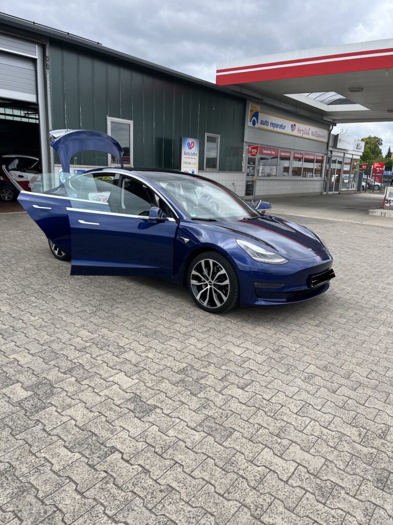 Eine blaue Tesla E Auto Limousine mit geöffneter Fahrertür und offenem Kofferraum steht vor einer Werkstatt in der Nähe einer Tankstelle in Deutschland.