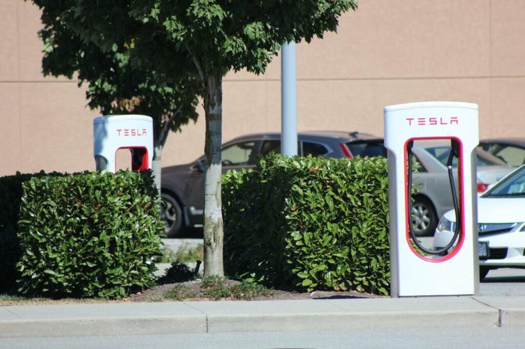 Zwei Tesla Supercharger Stationen für E Autos stehen auf einem Parkplatz in der Türkei, teilweise verdeckt durch Büsche, mit geparkten Autos und einem Baum in der Nähe.