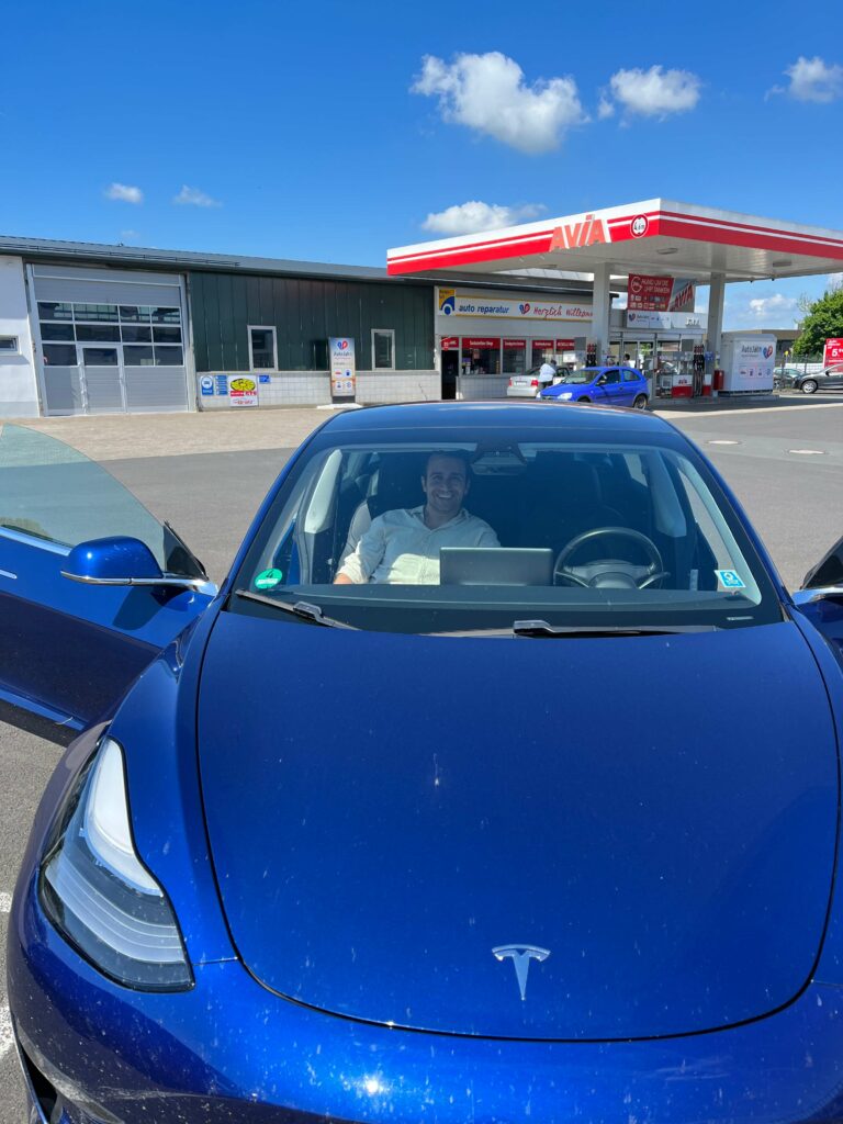 Eine Person sitzt in einem blauen E Auto Tesla, der an einem sonnigen Tag vor einer Tankstelle in Deutschland geparkt ist.