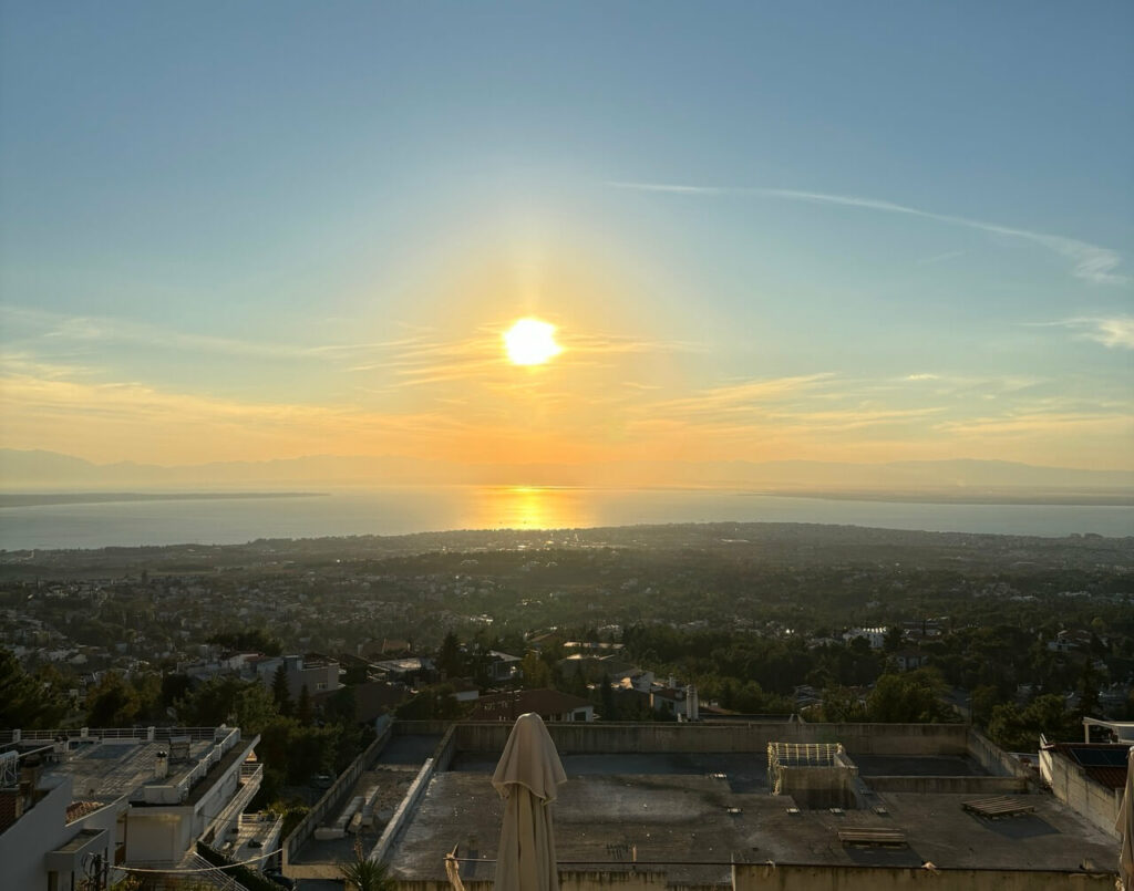 Blick auf die Stadt Thessaloniki bei Sonnenuntergang, wobei die tief stehende Sonne Spiegelungen auf dem Wasser erzeugt.