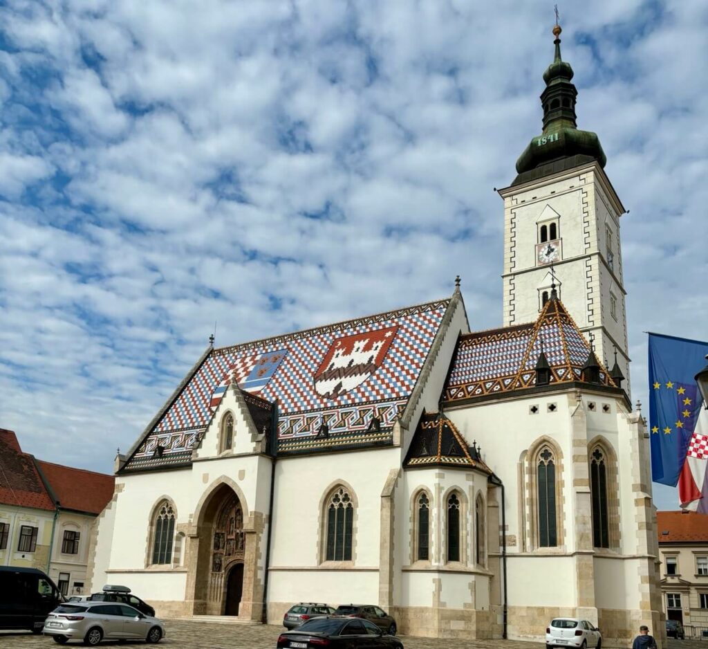 Die historische St.-Markus-Kirche in Zagreb mit einem bunten Ziegeldach mit Wappen, einem hohen Glockenturm unter einem teilweise bewölkten Himmel.